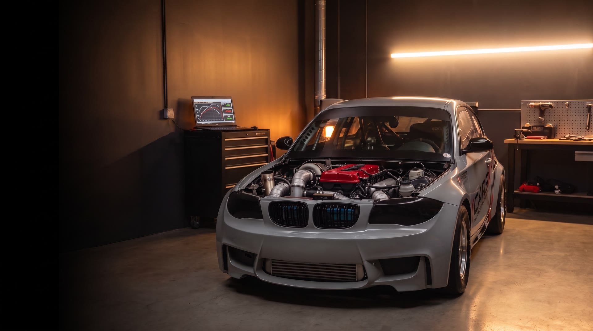 Mașina de drag E81 a CarsX în atelier, cu capota deschisă și capacul roșu de chiulasă luminat de o bandă LED caldă